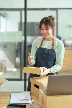 Küçük bir girişimin portresi, ve KME sahibi, Asyalı bir kadın girişimci, müşterinin iç kutusuna koymadan önce ürünü organize etmek için bir not defterine bilgi yazıyor..