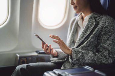 Başarılı bir Asyalı iş kadını ya da resmi takım elbiseli kadın girişimci bir uçakta birinci sınıf koltukta oturur ve uçuş sırasında akıllı telefon kullanır..