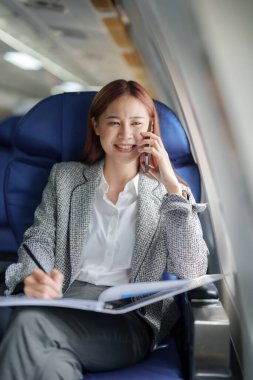 Başarılı bir Asyalı iş kadını ya da resmi takım elbiseli kadın girişimcinin portresi birinci sınıf koltukta oturuyor ve uçuş sırasında iş için akıllı telefon kullanıyor..