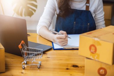 Girişimci, paket ürün kutusu hazırlamak için online alışveriş siparişlerini kontrol etmek için kalem noktası ve dizüstü bilgisayar kullanıyor. Müşteriye teslim edilecek mallar paketleniyor