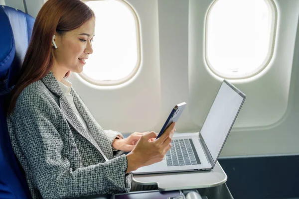 飛行機の中で正式なスーツを着て成功したアジアの実業家や女性起業家は ビジネスクラスの席に座って 飛行中にスマートフォンを使用します — ストック写真