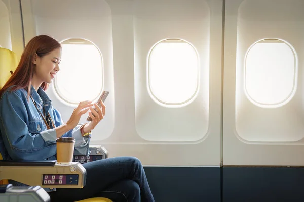 stock image Attractive portrait of Asian woman sitting at window seat in economy class using mobile phone during plane flight, travel concept, vacation.