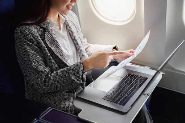 飛行機の中で正式な訴訟で成功したアジアの実業家や女性起業家は ビジネスクラスの座席に座っており 飛行中にコンピュータのラップトップを持つ文書を使用しています — ストック写真