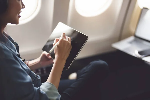 Asian woman sitting in a window seat in economy class using tablet and listening to instrumental music during an airplane flight, travel concept, vacation, relaxation.