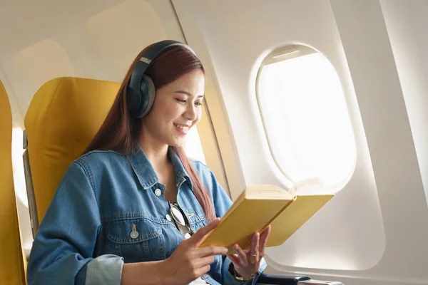 Stock image Attractive portrait of an Asian woman sitting in a window seat in economy class reading a book and listening to instrumental music during an airplane flight, travel concept, vacation, relaxation.