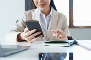 Sabahın erken saatlerinde masasında telefonunu, bilgisayarını ve mali belgelerini kullanırken gülümseyen bir Asyalı kadının portresi..