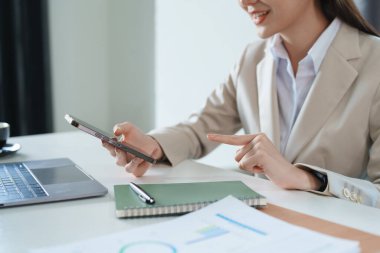 Sabahın erken saatlerinde masasında telefonunu, bilgisayarını ve mali belgelerini kullanırken gülümseyen bir Asyalı kadının portresi..