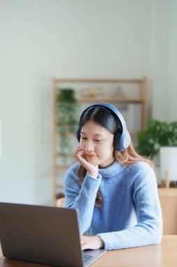 Bilgisayarı kullanan Asyalı genç bir kadının portresi, kulaklık takıp evdeki ahşap bir masada video konferansı yapıyor..