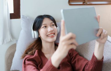 Portrait of asian woman using tablet and headphones relaxing on sofa at home.