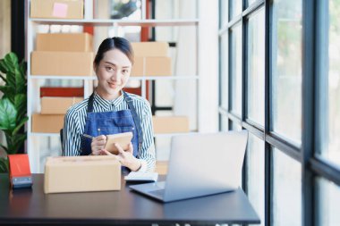 Bağımsız genç Asyalı kadın çevrimiçi satıcının küçük işletme girişimciliğine başlamak, bilgisayar kullanmak ve müşterilere dağıtım için ürünleri paketlemek için siparişler almaktır. KME dağıtım kavramı.