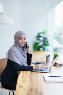 Güzel Müslüman kadın telefonda konuşuyor ve masasında bilgisayar kullanıyor..