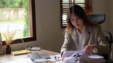 Asyalı genç bir iş kadını ya da muhasebeci. Bütçede çalışırken stresli yüzünü gösteriyor. Evrak işlerini yapıyor, bilgisayarını ahşap masaya koyuyor. Yüksek kalite 4k görüntü