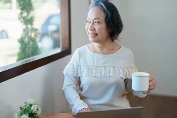 Portrait of an elderly Asian woman in modern pose doing computer work and drinking coffee