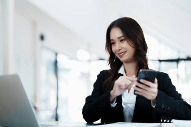 Sabahın erken saatlerinde masasında telefonunu, bilgisayarını ve mali belgelerini kullanırken gülümseyen bir Asyalı kadının portresi..