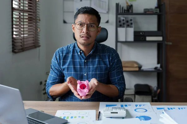 Portrait of an Asian male businessman showing a serious and anxious expression in an SME business , but no money in the pink pig piggy bank , saving plans