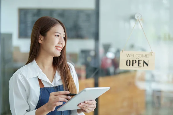 Portrait of a beautiful Asian woman running a small business holding a computer tablet showing a smiling face, a coffee shop owner opening a shop to welcome customers in the new morning, SME concepts.