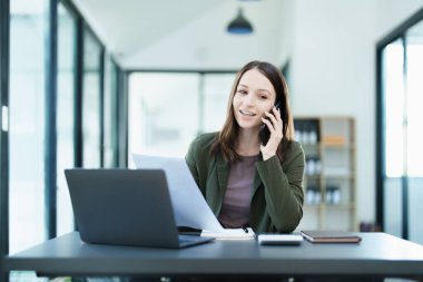 Portrait of a young Asian man showing a smiling face as she uses his phone, computer and financial documents on her desk in the early morning hours.