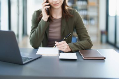 Sabahın erken saatlerinde masasında akıllı telefon, defter ve bilgisayarını kullanırken gülümseyen bir Asyalı kadının portresi.