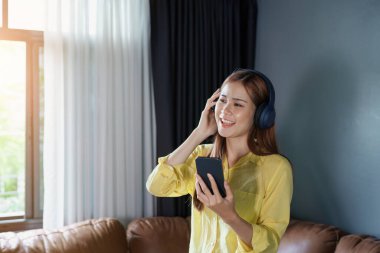 Asyalı kadın portresi akıllı telefon ve kulaklık kullanıyor evdeki kanepede dinleniyor..