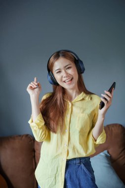 Asyalı kadın portresi akıllı telefon ve kulaklık kullanıyor evdeki kanepede dinleniyor..