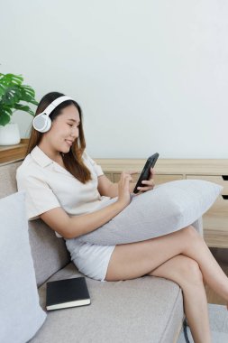 Asyalı kadın portresi akıllı telefon ve kulaklık kullanıyor evdeki kanepede dinleniyor..