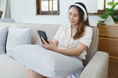 Asyalı kadın portresi akıllı telefon ve kulaklık kullanıyor evdeki kanepede dinleniyor..