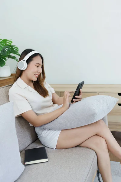 用智能手机和耳机在家里沙发上放松的亚洲女人的画像 — 图库照片
