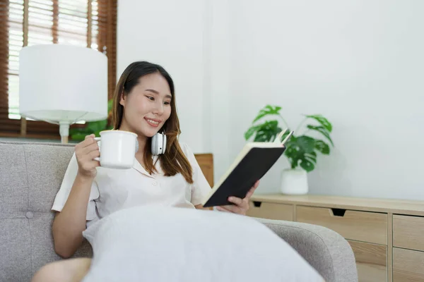 Ázsiai Olvasás Könyv Visel Fejhallgató Kávézni Csésze Pihentető Kanapén Otthon — Stock Fotó