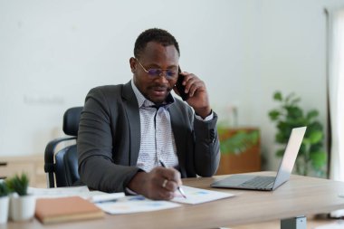 Orta yaşlı Amerikalı Afrikalı adam akıllı telefon ve dizüstü bilgisayar kullanarak finansal belge, vergi, döviz, muhasebe ve mali danışman üzerinde çalışıyor..