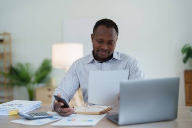 Orta yaşlı Amerikalı Afrikalı adam akıllı telefon ve dizüstü bilgisayar kullanarak finansal belge, vergi, döviz, muhasebe ve mali danışman üzerinde çalışıyor..