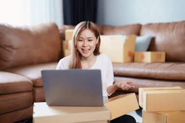 Bağımsız Asyalı kadın çevrimiçi satıcının küçük işletme girişimciliğini başlatarak bir müşteriyle bilgisayardan videoya konuşuyor ve müşteriye teslimat için ürünleri paketliyor. KME dağıtım kavramı.