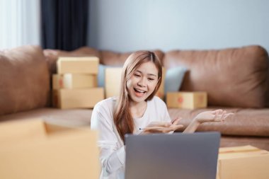 Bağımsız Asyalı kadın çevrimiçi satıcının küçük işletme girişimciliğini başlatarak bir müşteriyle bilgisayardan videoya konuşuyor ve müşteriye teslimat için ürünleri paketliyor. KME dağıtım kavramı.