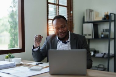 Orta yaşlı Amerikalı Afrikalı iş adamı bilgisayarlı dizüstü bilgisayarı elinde tutuyor ve kazanan elini kaldırıyor. Başarı başarısını kutlamaktan mutluluk duyuyoruz..