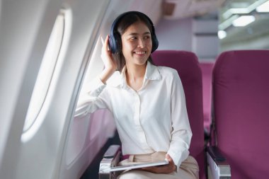 Genç Asyalı çekici iş kadını yolcu rahat olmak için kablosuz kulaklık takıyor ve uçuş sırasında tabletle çalışıyor..