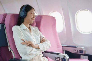 Genç Asyalı çekici iş kadını yolcu rahat olmak için kablosuz kulaklık takıyor ve uçuş sırasında tabletle çalışıyor..