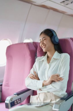 Genç Asyalı çekici iş kadını yolcu rahat olmak için kablosuz kulaklık takıyor ve uçuş sırasında tabletle çalışıyor..