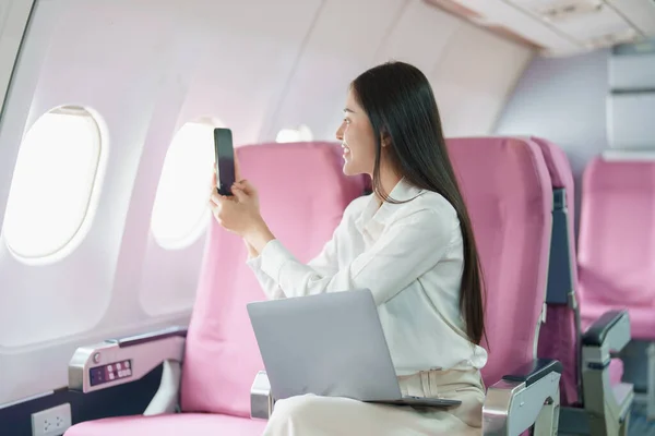 Young Asian attractive business woman passenger sitting on business class luxury plane while using smart phone mobile to take a photo and use computer laptop during the flight.