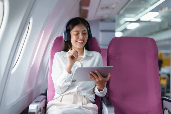 Young Asian attractive business woman passenger sitting on business class luxury plane while wearing wireless headphone for relax and working with tablet during the flight.