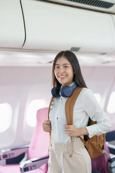 stock image Young asian attractive woman travel by airplane, Passenger wearing headphone putting hand baggage in lockers above seats of plane.