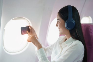 Genç Asyalı çekici iş kadını yolcu kablosuz kulaklık takıp birinci sınıf lüks uçakta oturuyor ve uçuş sırasında rahatlamak için akıllı telefon kullanıyor..