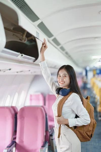 stock image Young asian attractive woman travel by airplane, Passenger wearing headphone putting hand baggage in lockers above seats of plane.