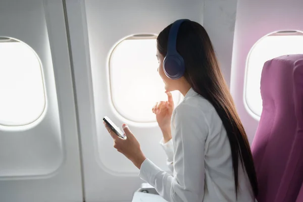 Young Asian attractive business woman passenger sitting on business class luxury plane while wearing wireless headphone and using smart phone mobile for relax during the flight.