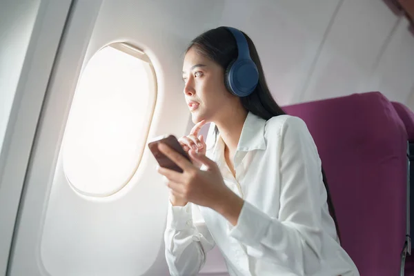 stock image Young Asian attractive business woman passenger sitting on business class luxury plane while wearing wireless headphone and using smart phone mobile for relax during the flight.