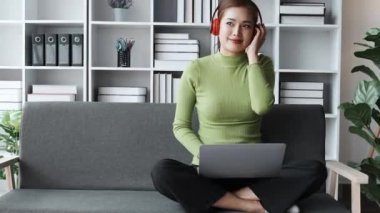 Genç Asyalı kadın kulaklık takıyor gülen yüzünü gösteriyor ve evdeki kanepede dinlenirken müzik dinlemek için bilgisayar kullanıyor. Yüksek kalite 4k görüntü