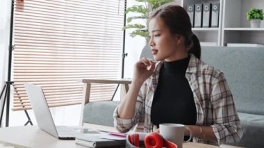 Evde çalışan Asyalı genç kadın, gülümseyerek ve evde bilgisayar kullanarak düşünceli bir jest yaparken belgeleri kontrol ediyor. Yüksek kalite 4k görüntü