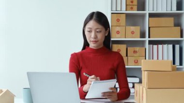 e-ticaret, küçük işletme, sme, güzel Asyalı kadın bilgisayara bakıyor ve not alıyor müşteri siparişleri alıyor ve çalışırken gülen yüzle çevrimiçi satışlardan para kazanıyor. Yüksek kalite 4k