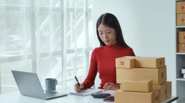 E-ticaret, küçük işletme, sme, nota bakan güzel Asyalı kadın ve hesap makinesi kullanarak müşteri siparişlerini hesaplıyor ve çalışırken gülen yüzle çevrimiçi satışlardan para kazanıyor.