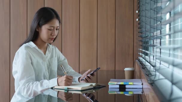 Młoda Kobieta Azjatka Pracuje Smartfonie Komórkowym Robi Notatki Notatniku Ekspresją — Wideo stockowe