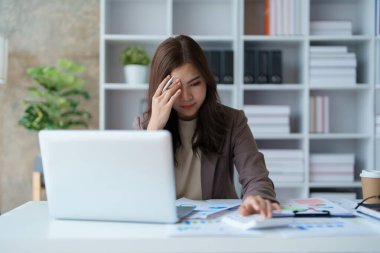 Sabahın erken saatlerinde masasında hesap makinesi, finansal döküman ve bilgisayar bilgisayarını kullanırken ciddi bir yüz ifadesi gösteren Asyalı genç bir kadının portresi.