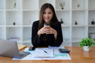 İşyerinde bilgisayarları ve finansal bütçe belgelerini kullanarak işini başarıyla yürüttüğü için gülümseyen bir kadın iş sahibinin portresi.